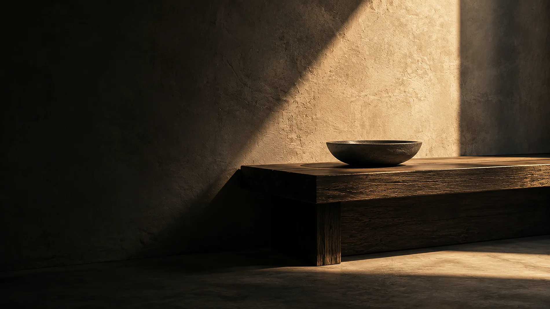A low oak bench and a shallow stone bowl against charcoal plaster, crossed by a band of late-afternoon light.