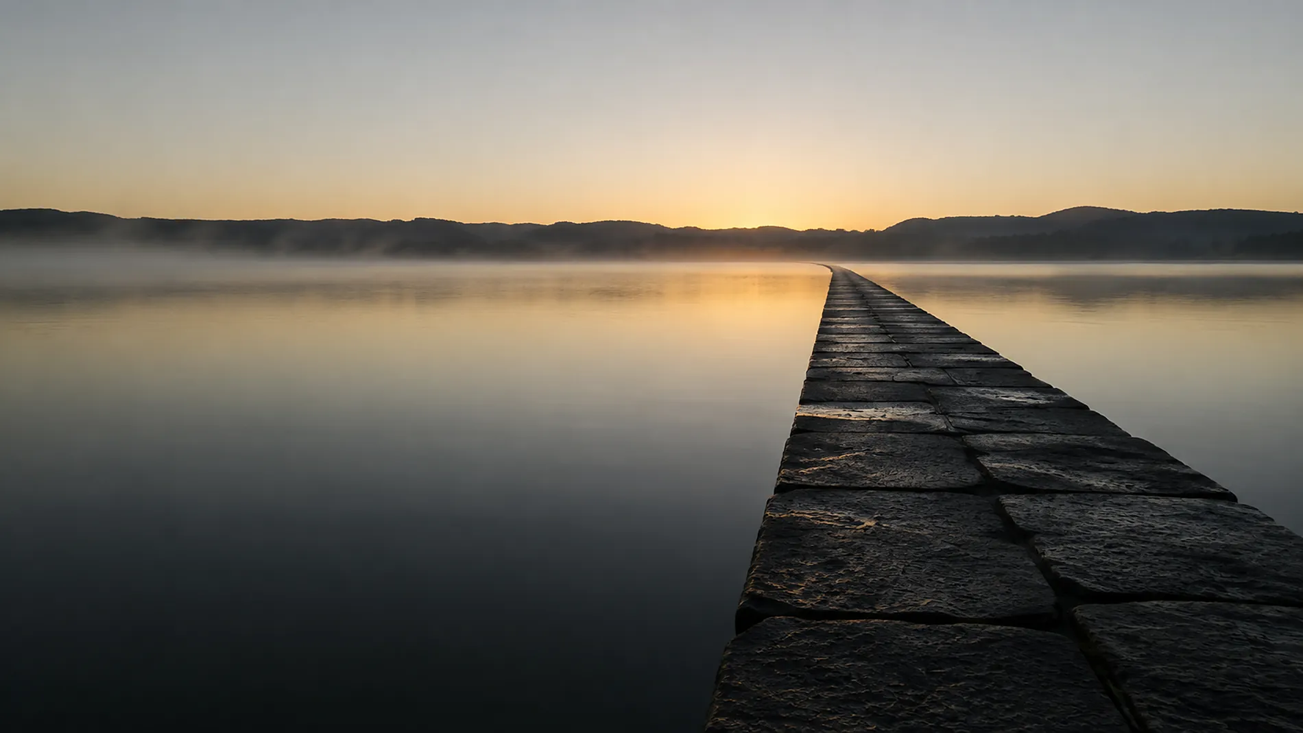 A low stone causeway running across still water toward a distant ridge at first light.